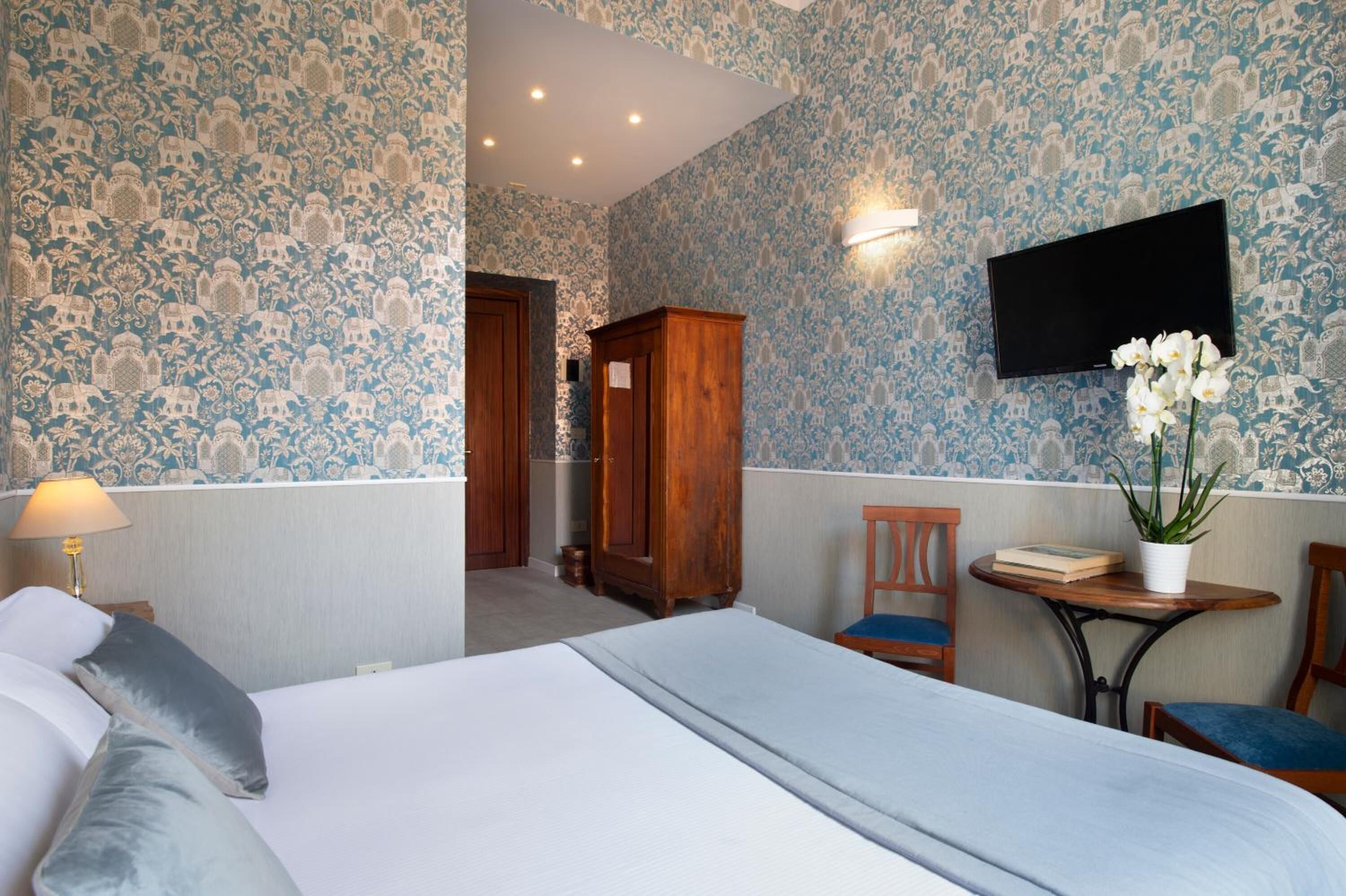 A hotel room with a bed, a desk with a chair, a TV mounted on the wall, and a wooden wardrobe.