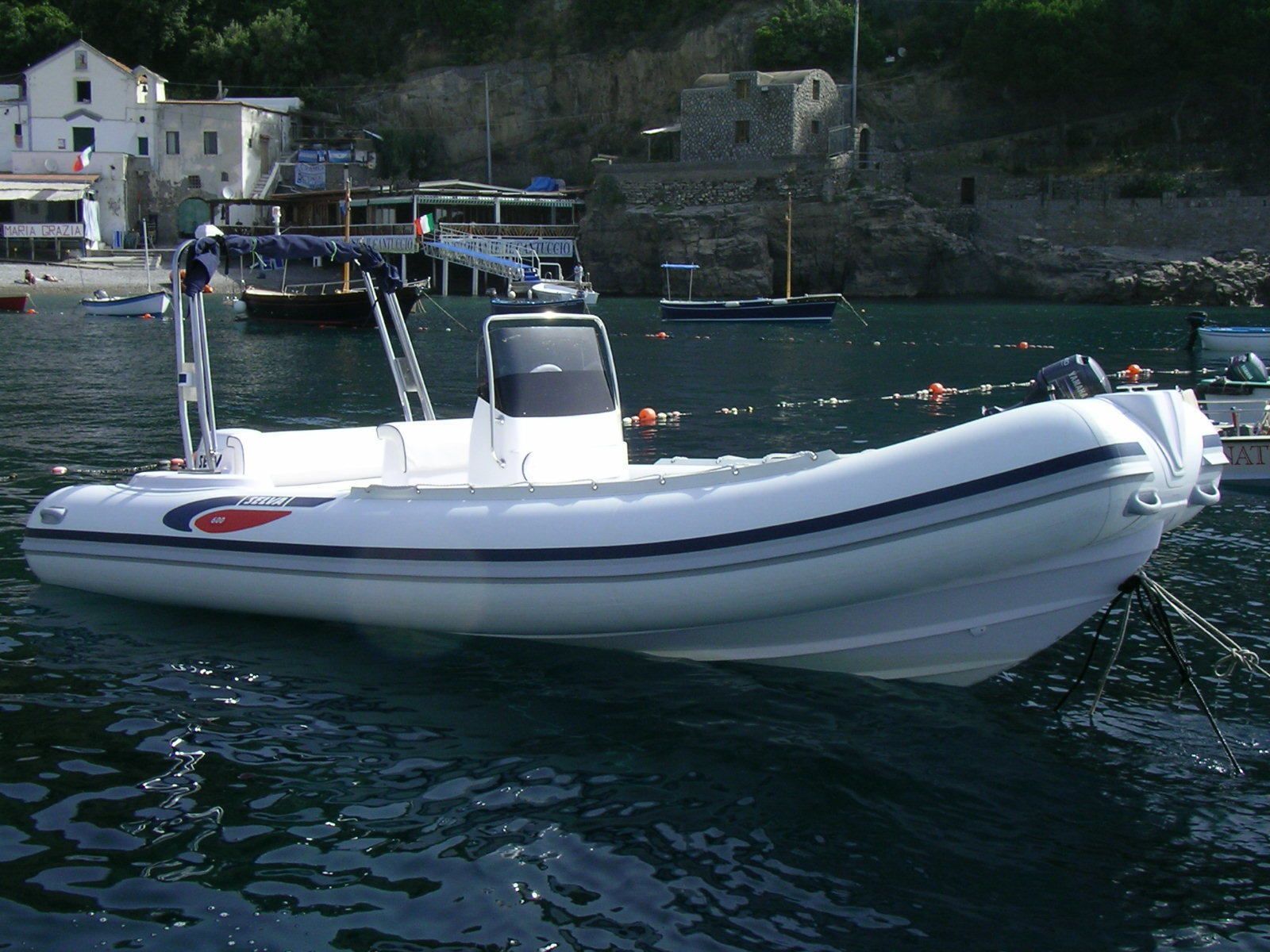 Ein kleines, weißes Motorboot liegt in einem Hafen vor einer Küste mit steilen Klippen und Gebäuden im Hintergrund.