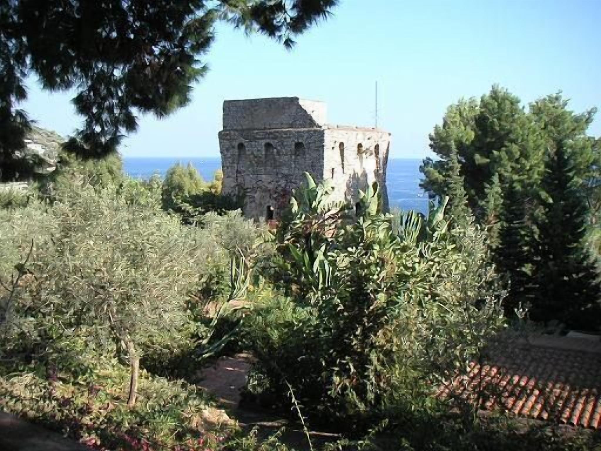 A stone building with arched windows, surrounded by greenery and overlooking a body of water.