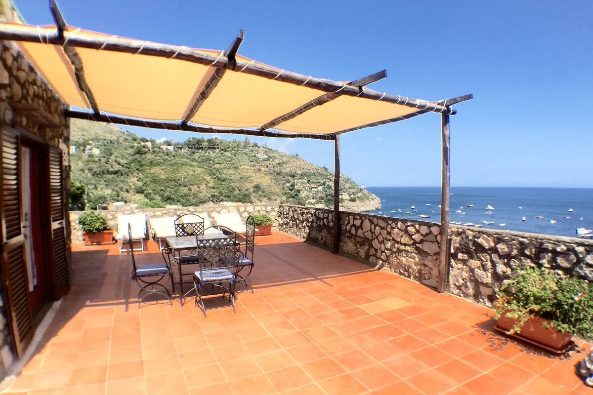 A terrace with a table and chairs, a stone wall, and a view of the sea.