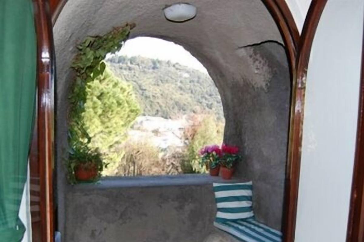 A narrow balcony with a view of a mountainous landscape, a small seating area with a striped cushion, and potted plants.