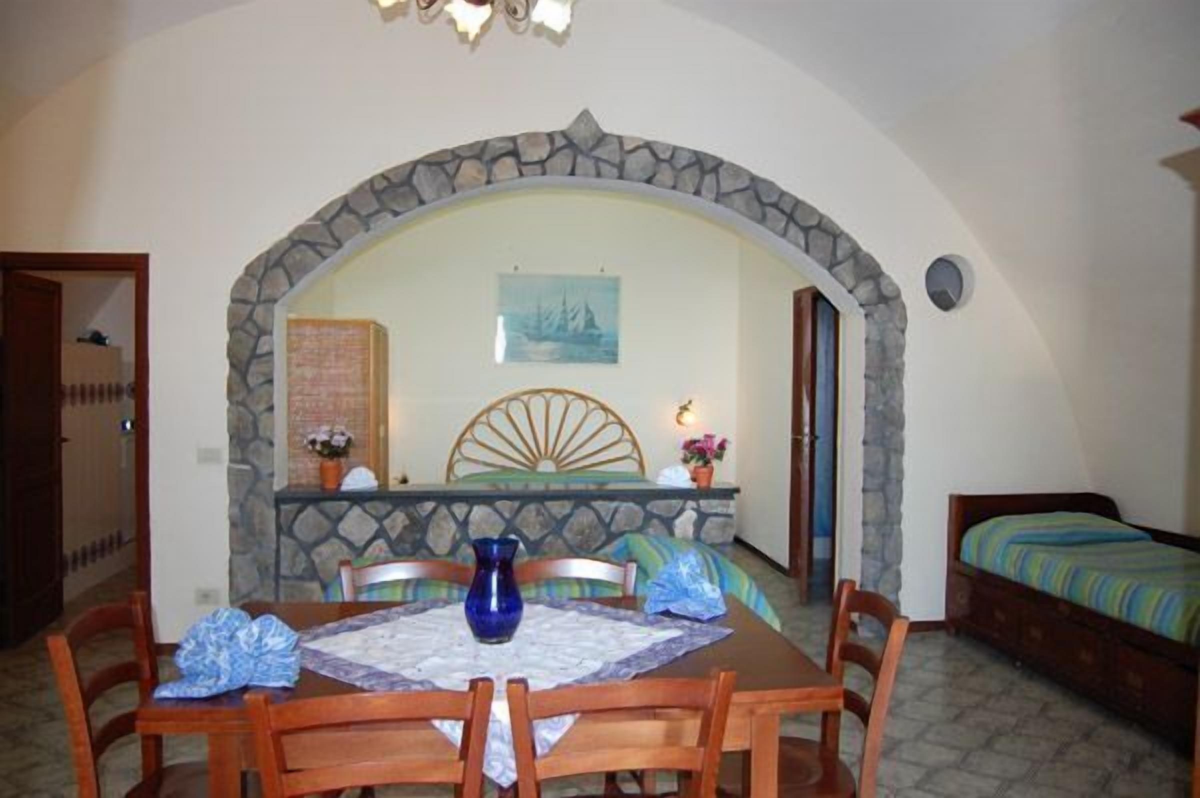 Un coin repas agrémenté d’une arche en pierre, une table avec des chaises, un vase et un lit recouvert d’une courtepointe à rayures.
