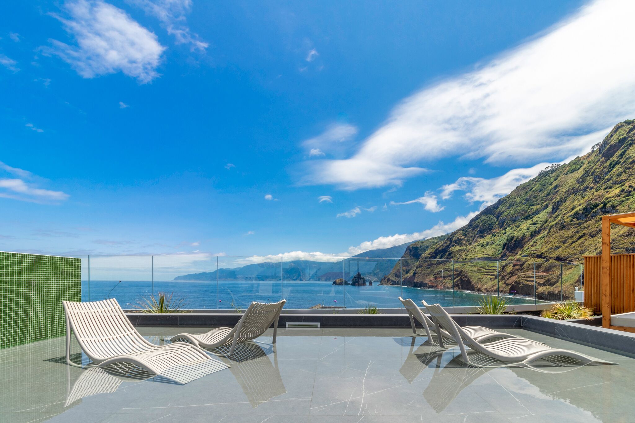 A rooftop infinity pool with lounge chairs, overlooking a coastal landscape with mountains and the sea.
