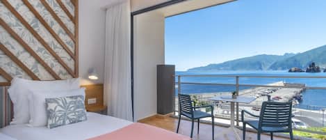 A hotel room with a large bed, a view of the sea, and a balcony with chairs and a table.