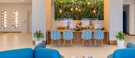 Un espace d’accueil moderne avec un comptoir de réception en bois, des chaises à motifs et un mur végétal agrémenté de luminaires suspendus.
