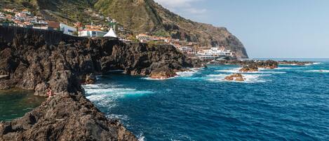 A coastal scene with a person swimming in clear water, rocky cliffs, and a building complex in the background.