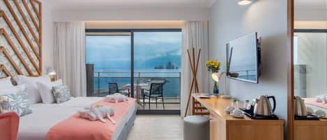 A hotel room with a bed, a desk, a chair, a TV, and a balcony with a view of the sea.
