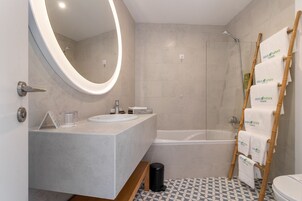 Un baño moderno con un espejo redondo, una bañera, una ducha y un inodoro. Hay un perchero con toallas y un cesto de basura.