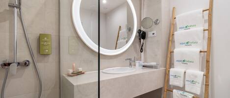 Un baño moderno con ducha de vidrio, un espejo redondo, tocador con lavamanos y un perchero de madera con toallas.