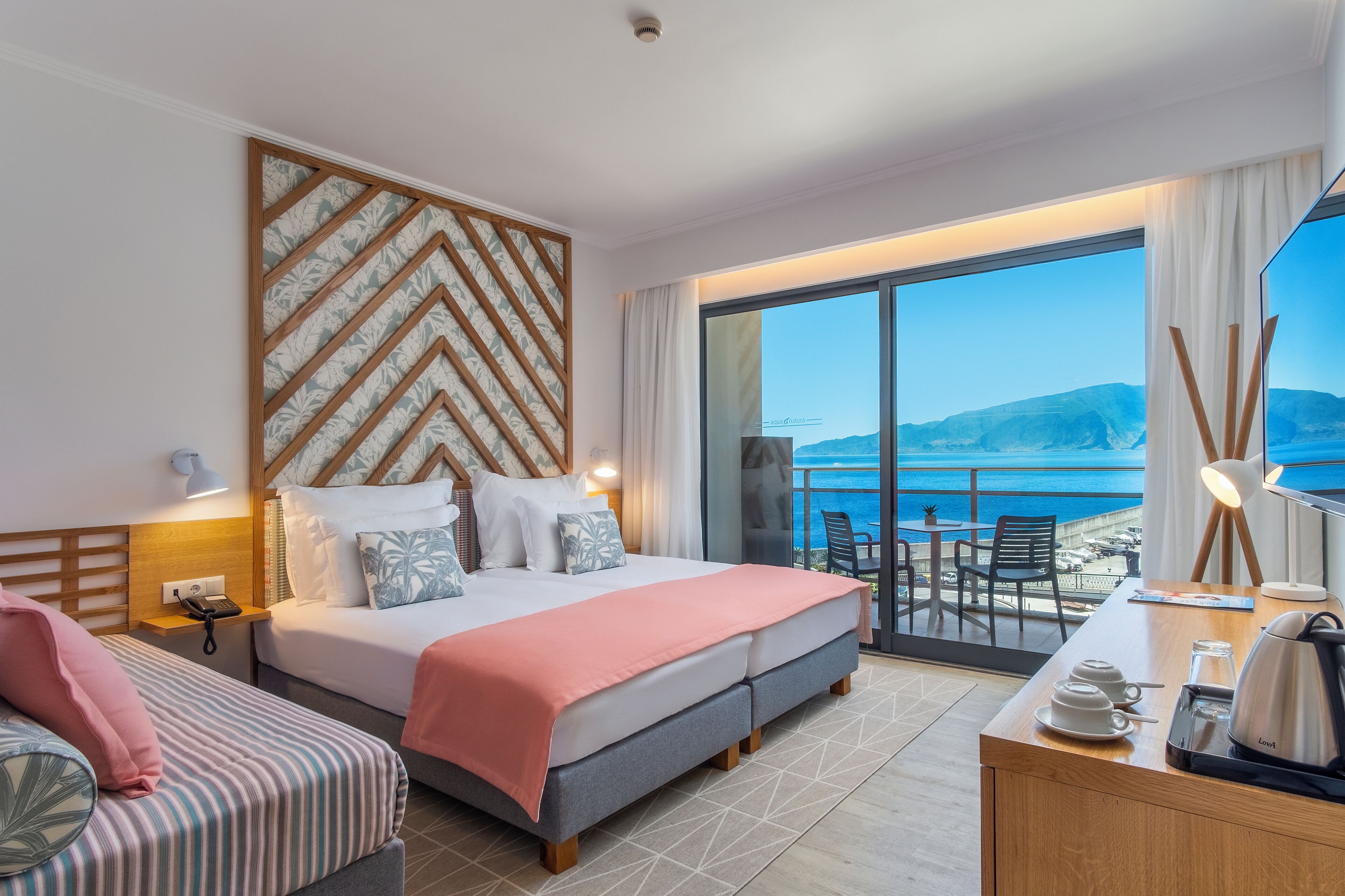 A hotel room with a bed, a desk, a chair, a small table, and a view of the sea.
