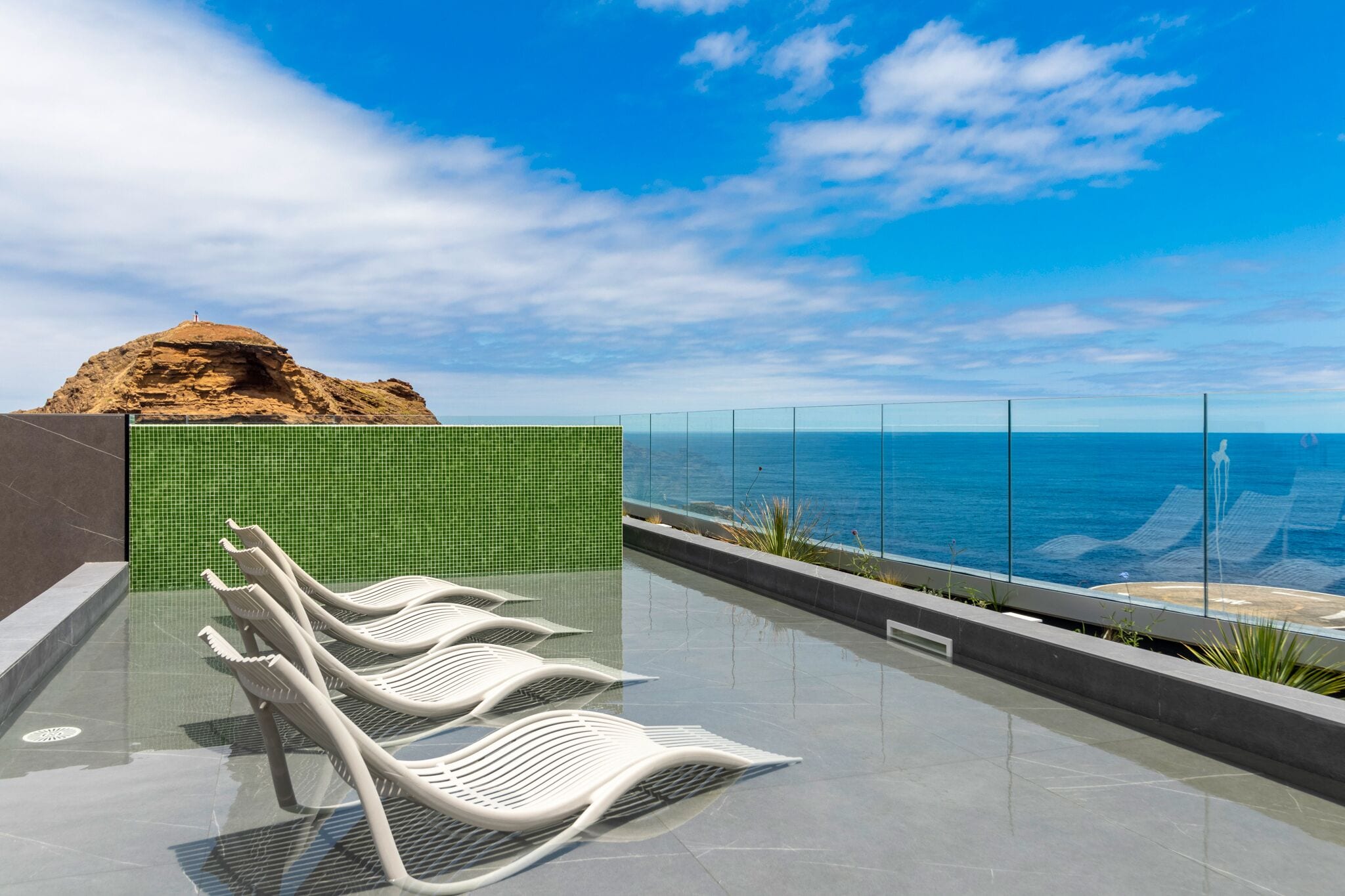 A pool area with white loungers, a green tiled wall, and a view of the ocean.
