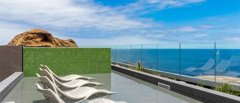 A pool area with white loungers, a green tiled wall, and a view of the ocean.