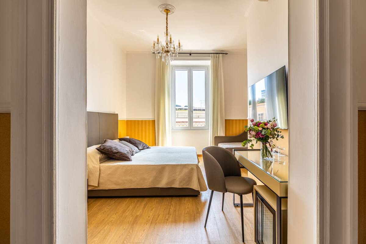 Una camera d'albergo con un letto, una scrivania, una sedia, una TV e una finestra con tende.