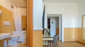 Un baño con inodoro, lavamanos y ducha. Hay un espejo y una mesita con un florero.