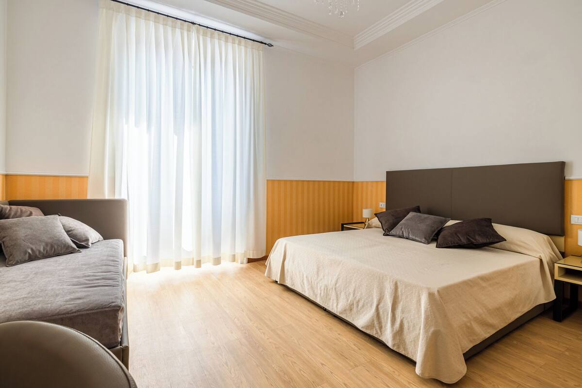 Una camera d'albergo con un letto grande, un divano più piccolo, un comodino e una finestra con tende leggere.