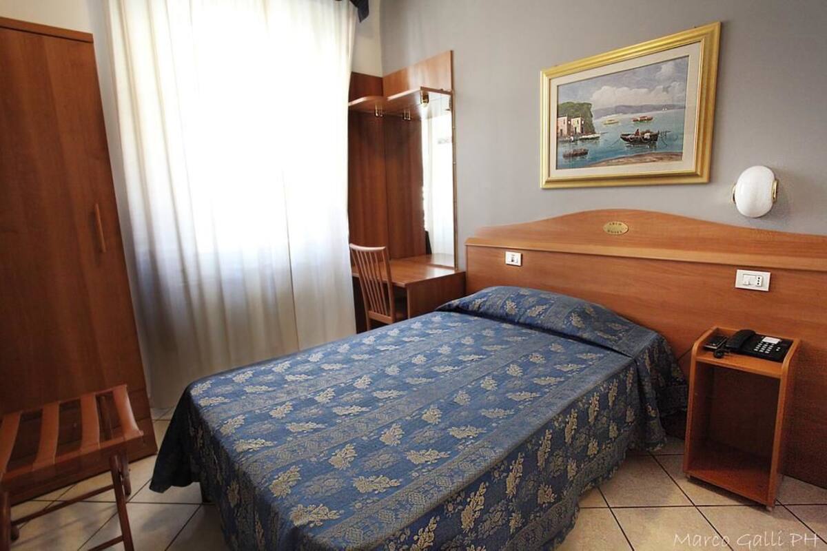 Une chambre d’hôtel avec un lit, un bureau, une chaise, une armoire et un tableau au mur.