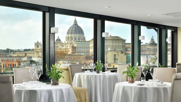 A dining area with tables set for two, large windows offering a cityscape view, and a prominent dome building in the background. - Orange Garden Bed & Breakfast (Rome)
