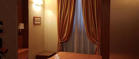 Habitación de hotel con cama, escritorio, silla, lámpara y ventana con cortinas.