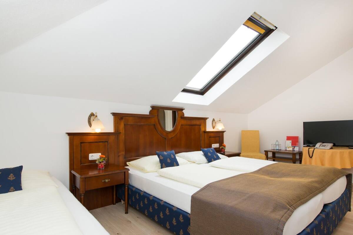 A hotel room with a skylight, a large bed with a wooden headboard, a bedside table with a lamp, a chair, and a desk with a telephone.