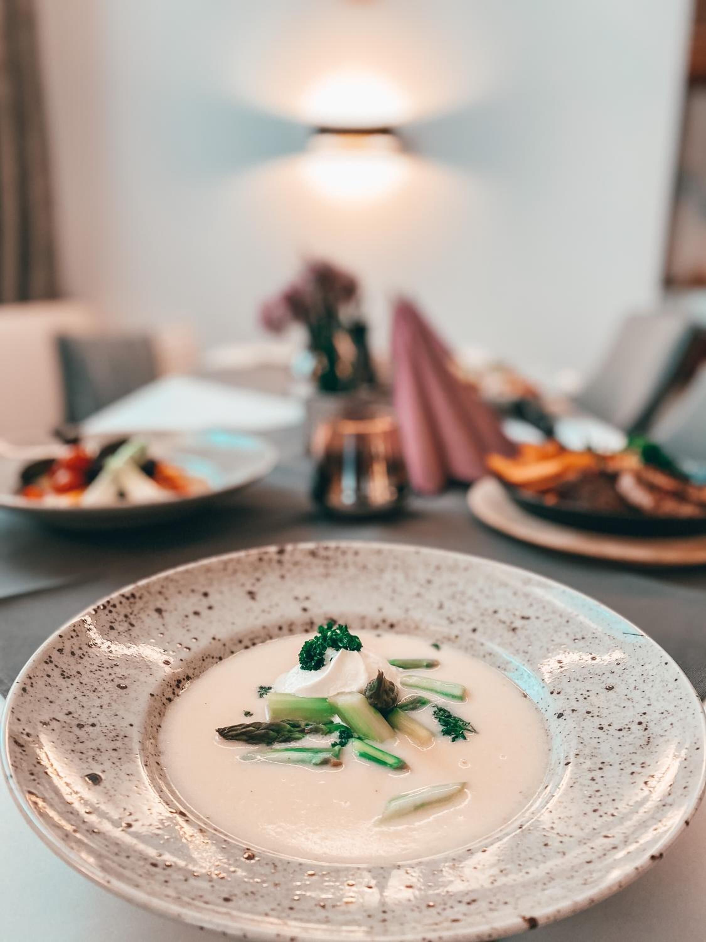A dining table with a plate of creamy soup garnished with herbs, a candle, and a vase with flowers in the background.