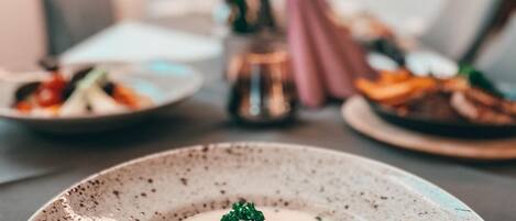 A dining table with a plate of creamy soup garnished with herbs, a candle, and a vase with flowers in the background.