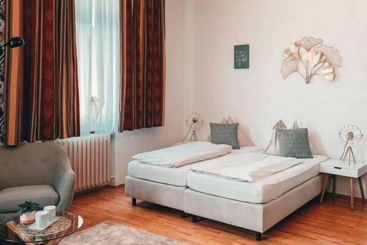 A bedroom with a double bed, a small side table, a chair, a radiator, and a window with curtains.