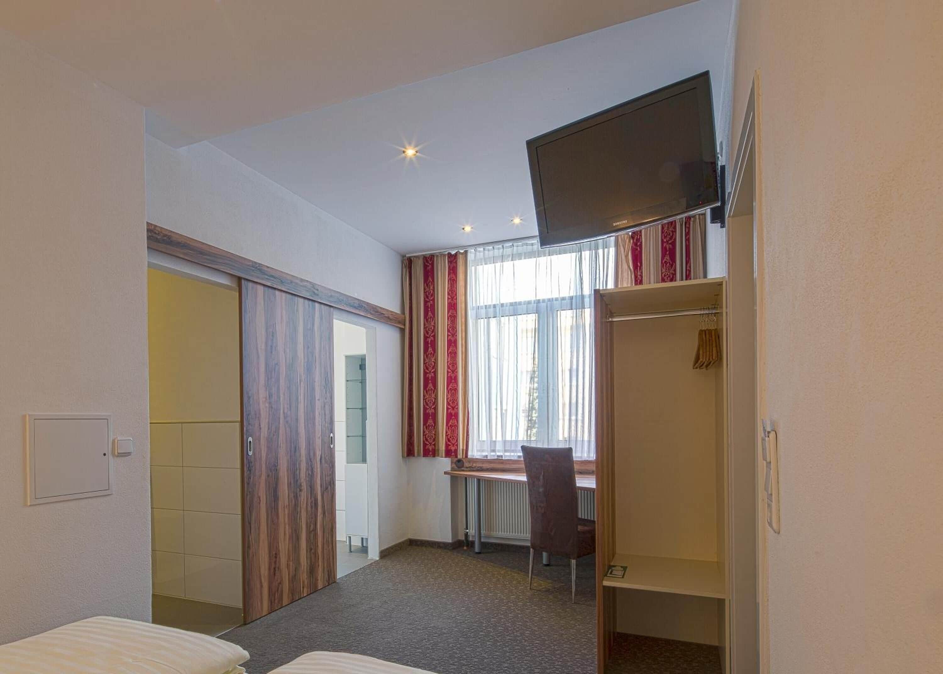 A hotel room with a bed, a desk, a chair, a wardrobe, and a television.
