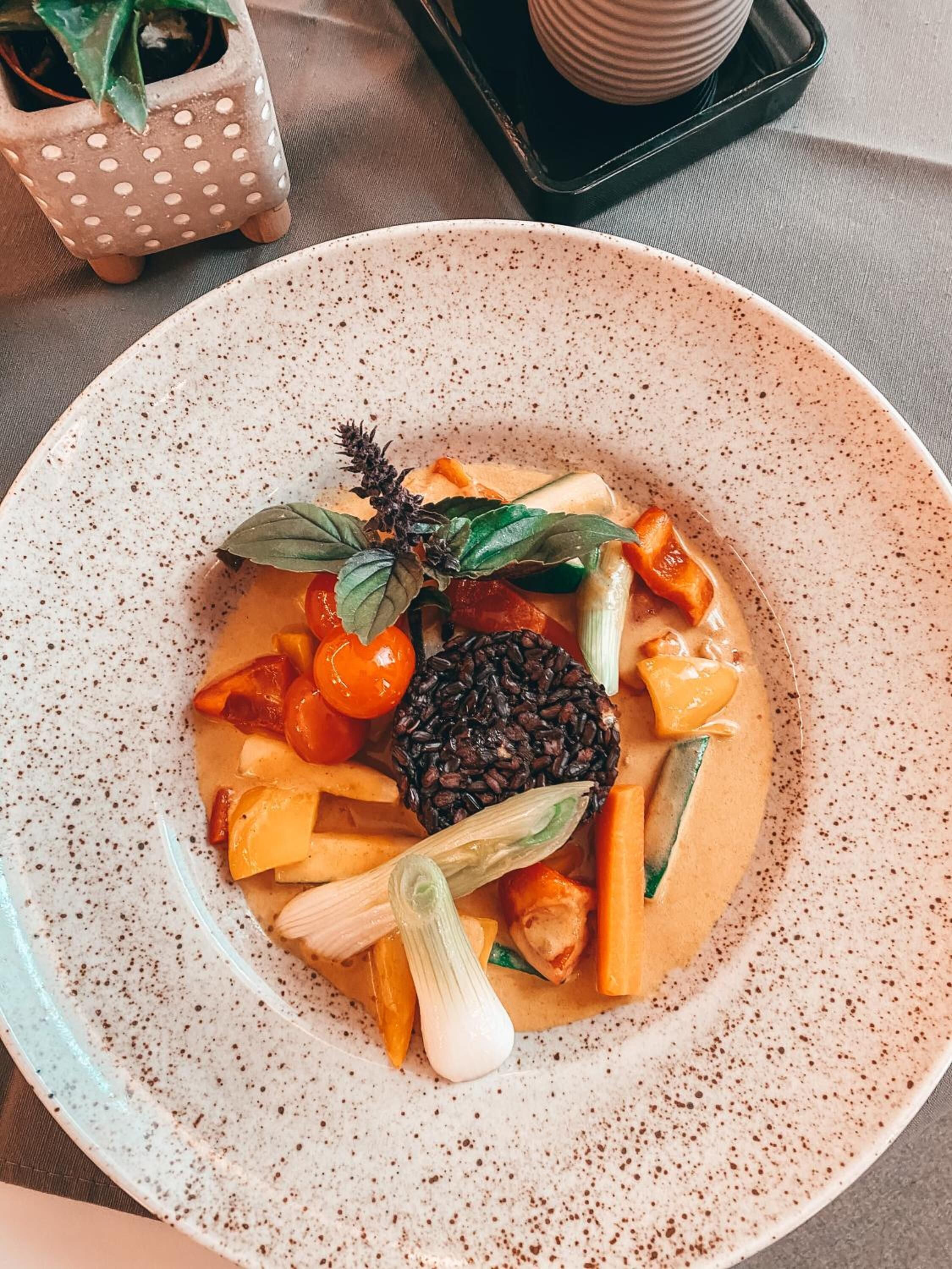 A bowl of food with various vegetables and a grain-like item, possibly rice, on a speckled ceramic plate.