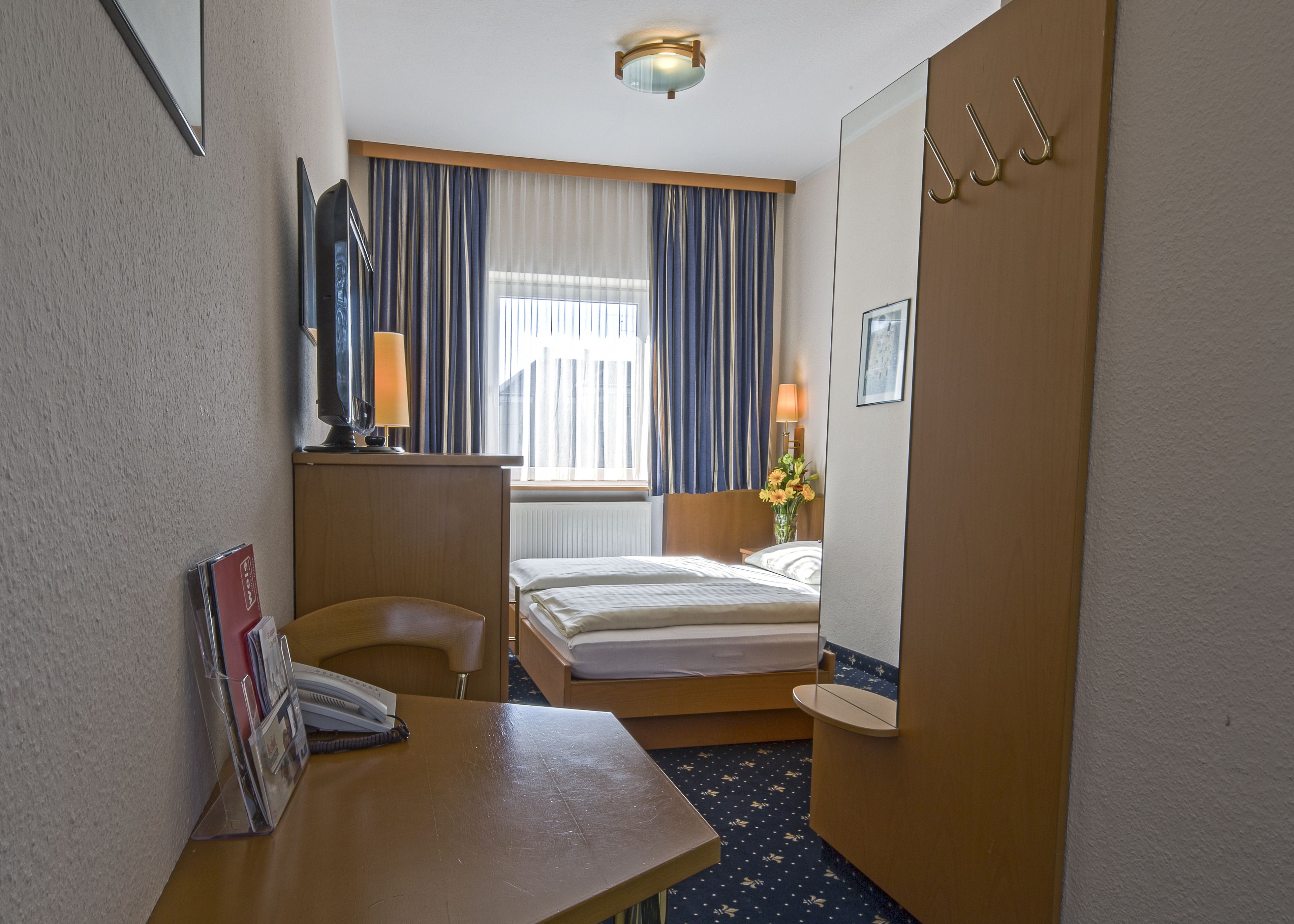 Ein Hotelzimmer mit einem Bett, einem Schreibtisch mit Lampe, einem Sessel, einem Fenster mit Vorhängen und einem Spiegel.
