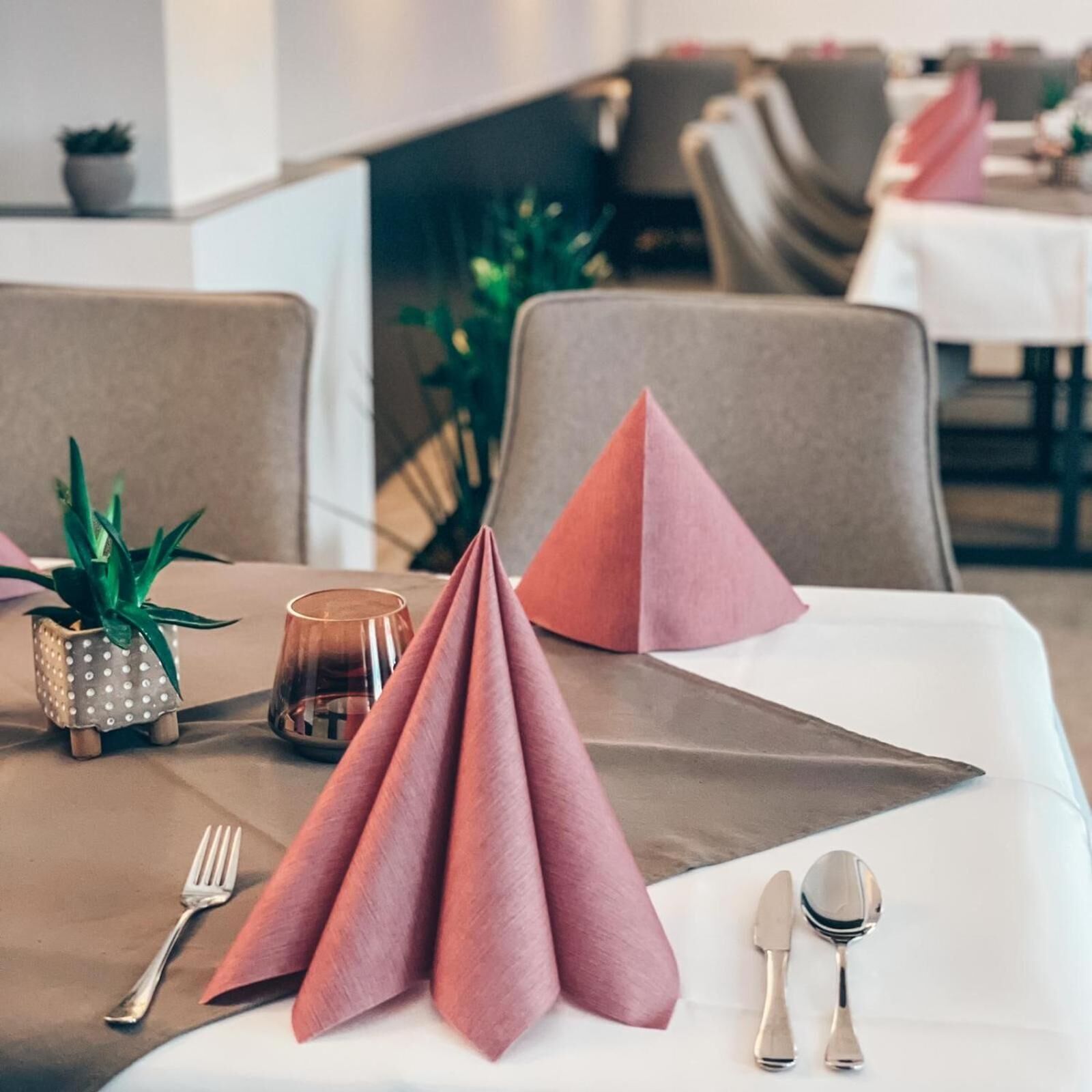 A dining table with a white tablecloth, pink napkins, a small potted plant, a candle, and cutlery.