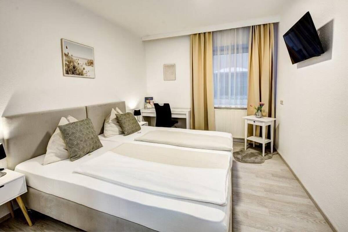 A hotel room with two beds, a desk, a chair, a TV mounted on the wall, and a window with curtains.