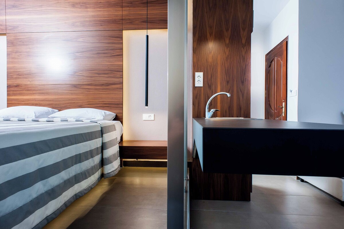 A modern hotel room with a wooden headboard, a bed with striped bedding, a bedside table, and a built-in wooden vanity with a sink.