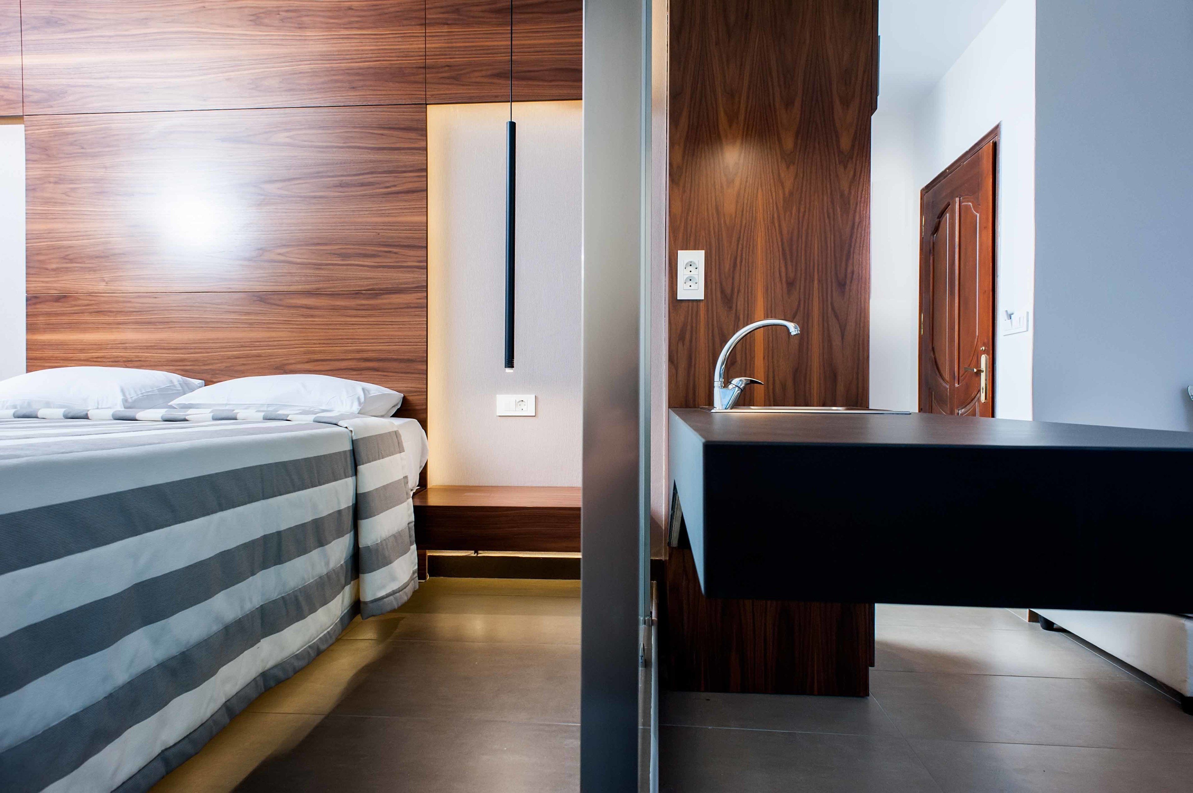 A modern hotel room with a wooden headboard, a bed with striped bedding, a bedside table, and a built-in wooden vanity with a sink.