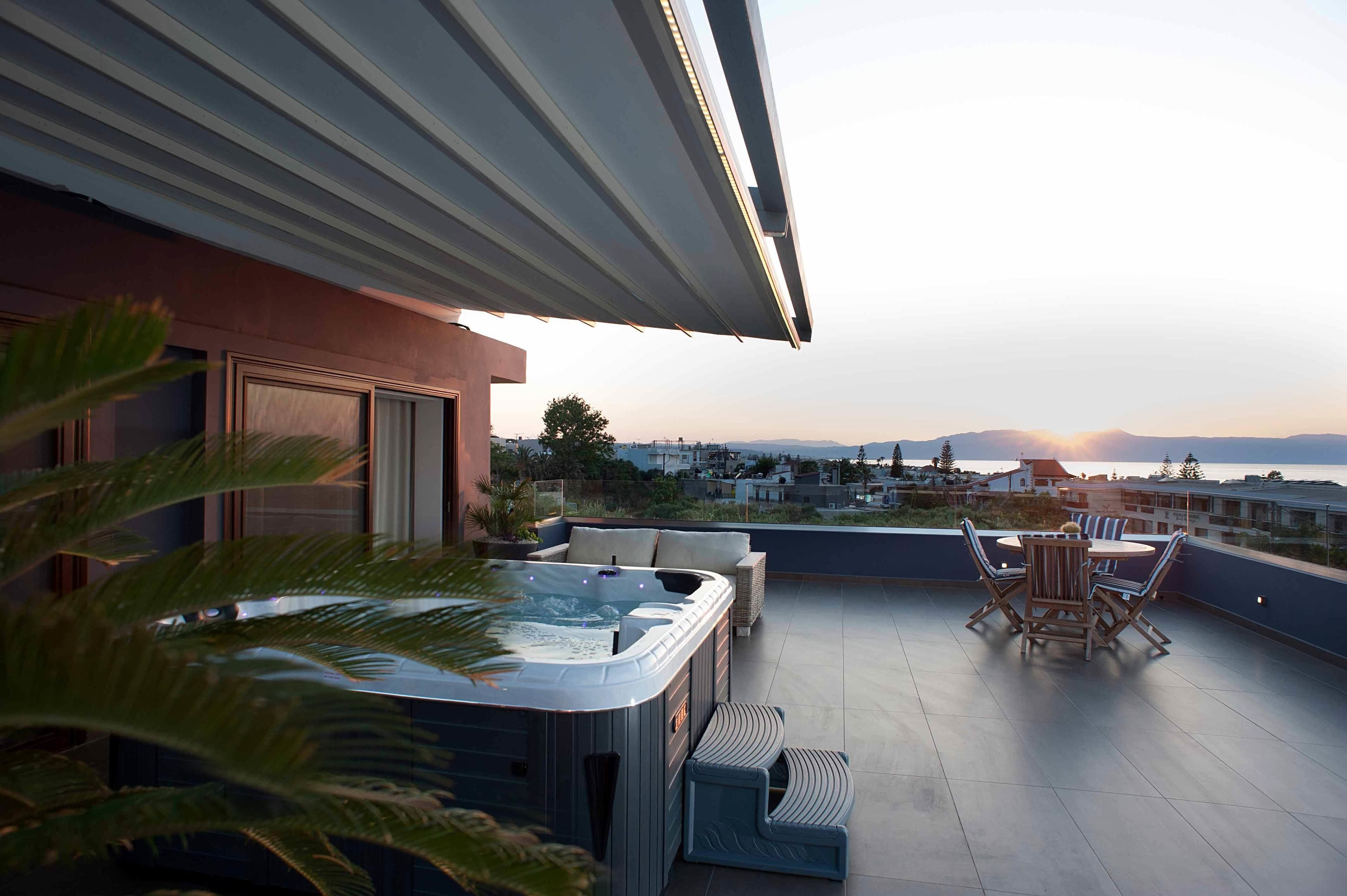 A rooftop terrace with a hot tub, outdoor seating, and a view of the cityscape and ocean at sunset.
