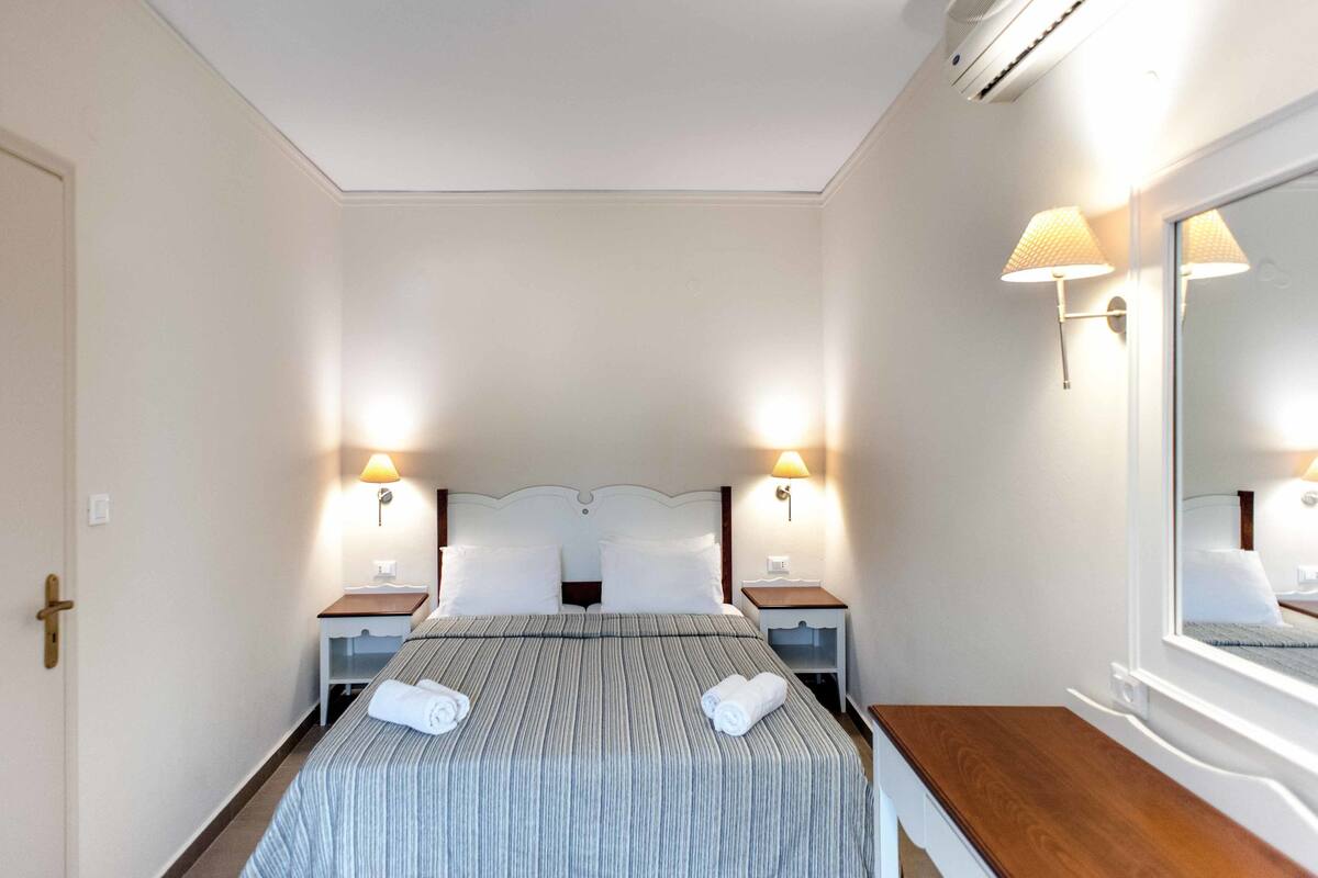 A neatly made bed with two pillows and two rolled towels, a bedside table with a lamp, and a large mirror on the wall.