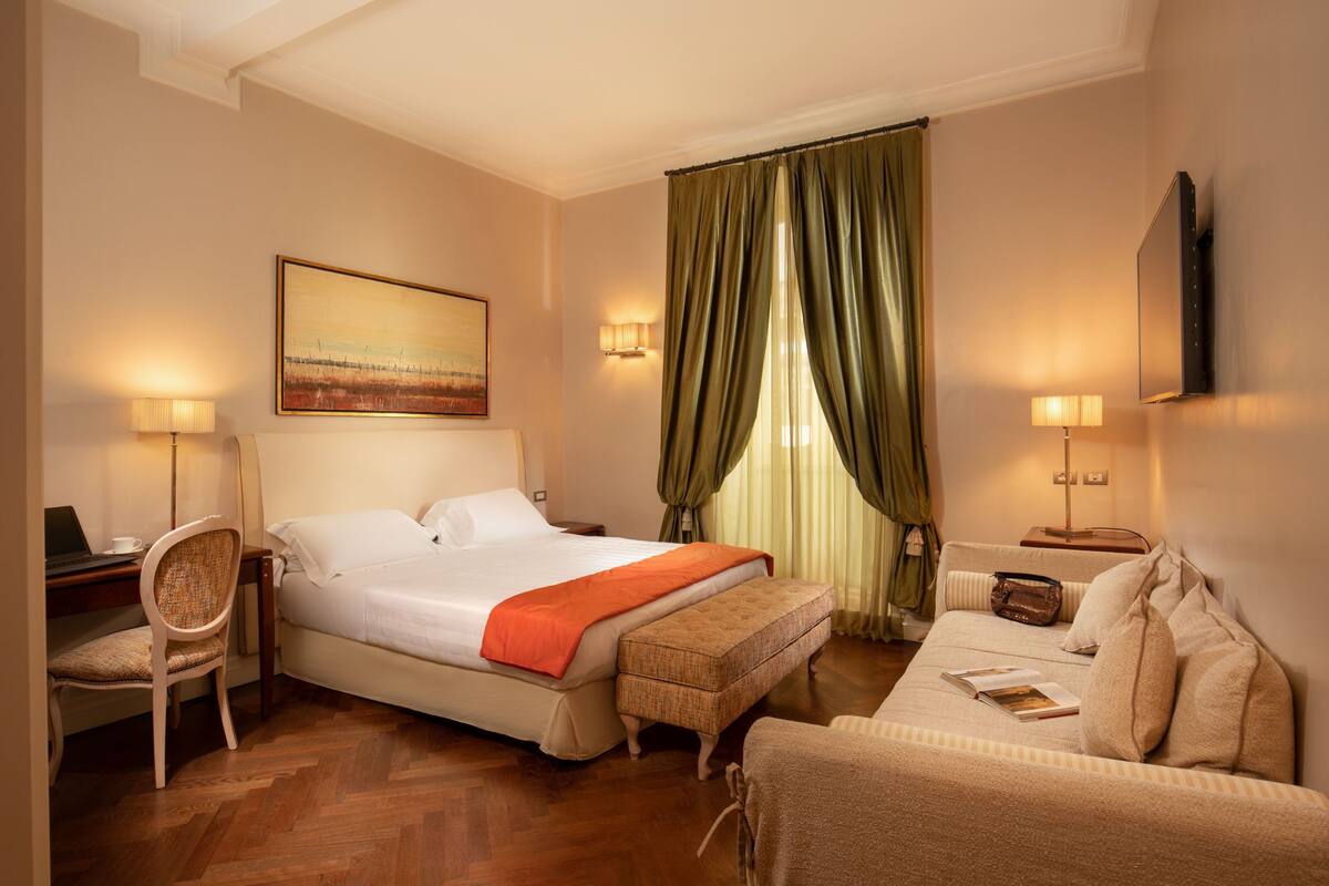 A hotel room with a bed, a desk with a laptop, a chair, a sofa, and a television on the wall.