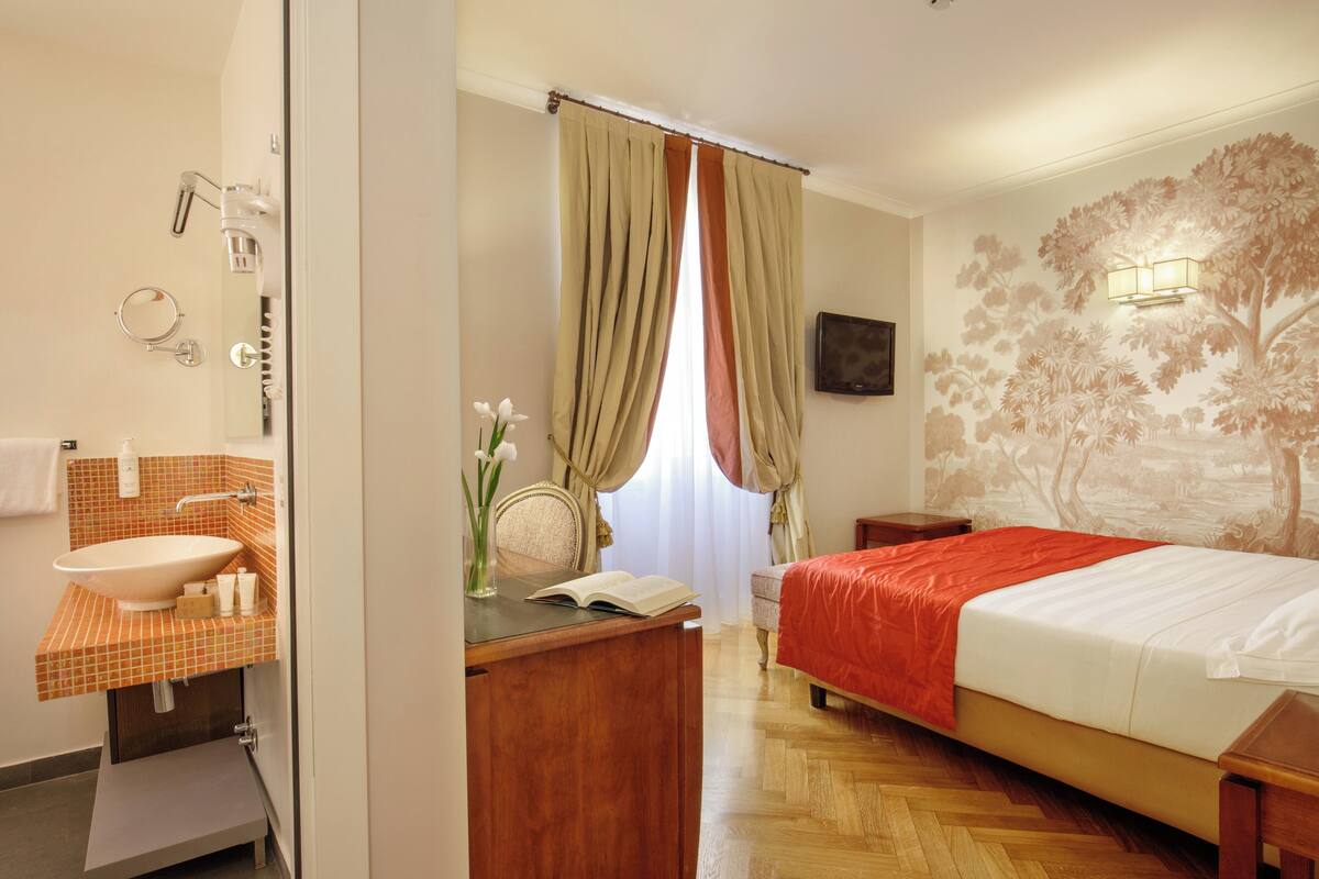 A hotel room with a bathroom, a bed, a desk, and a window with curtains.