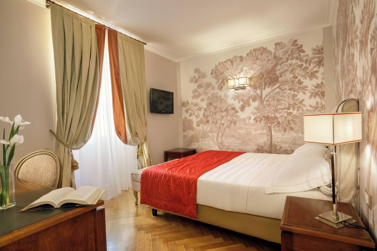 A hotel room with a bed, a bedside table, a lamp, a chair, a desk with a book, and a window with curtains.