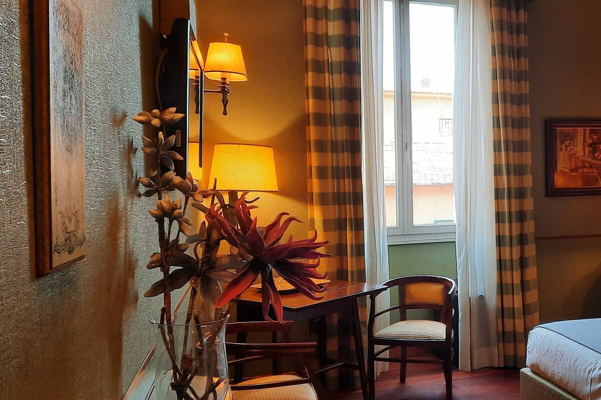 Habitació d'hotel amb un llit, una escriptori amb una cadira, una làmpada, una finestra amb cortines i un quadre penjat a la paret.