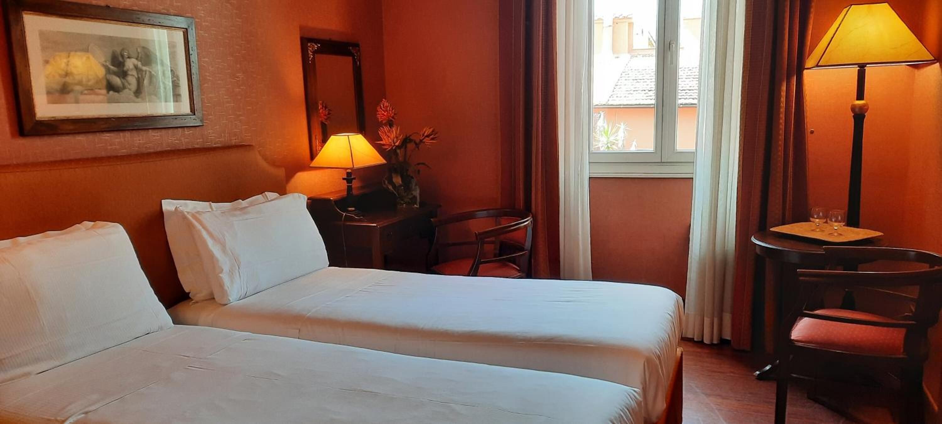 A hotel room with two beds, a desk with a lamp, a chair, a mirror, and a window with curtains.