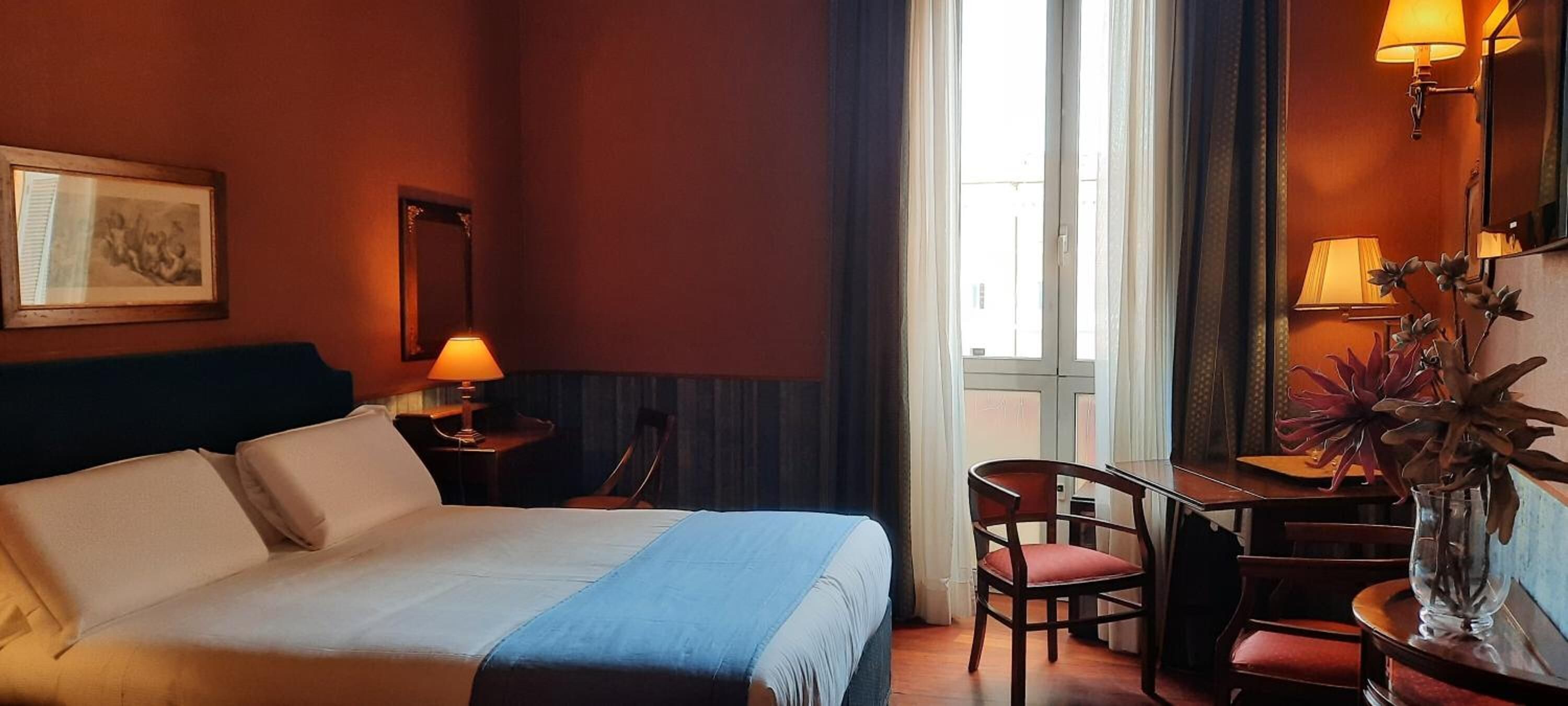 Quarto de hotel com uma cama, uma escrivaninha e uma cadeira. Há uma janela com cortinas, uma luminária na escrivaninha e uma planta na mesinha de apoio.