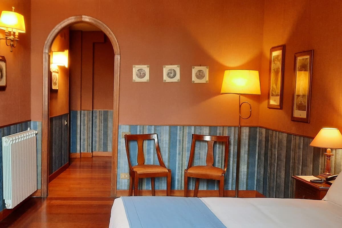 Habitació d'hotel amb un llit, dues cadires, un vas amb flors, una làmpada i un mirall.