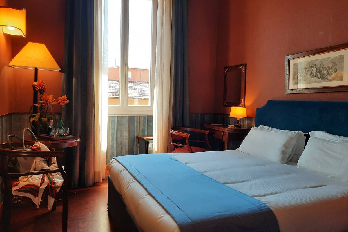 Una habitació d'hotel amb llit, escriptori, cadira, làmpada, finestra i un quadre al mur.