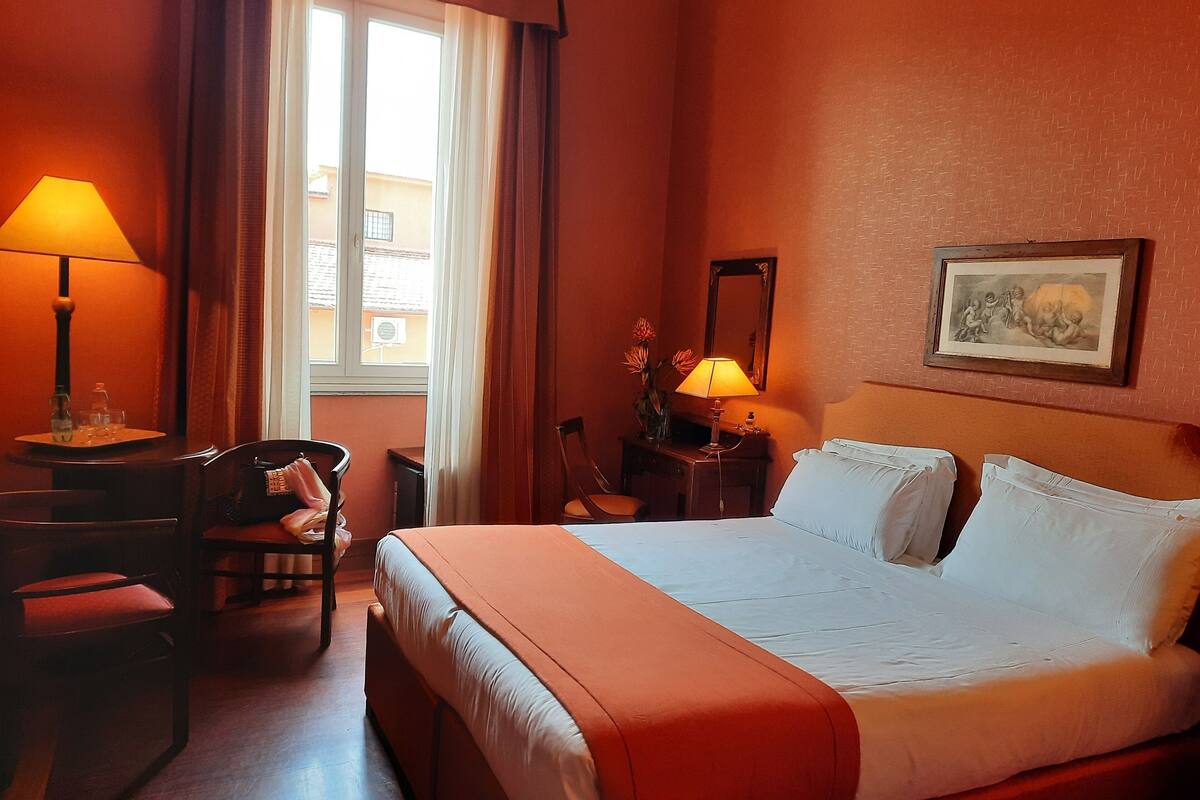 Habitació d'hotel amb un llit, una taula de treball amb una làmpada, una cadira, una nitzeta amb una làmpada, un mirall i un quadre amb marc a la paret.