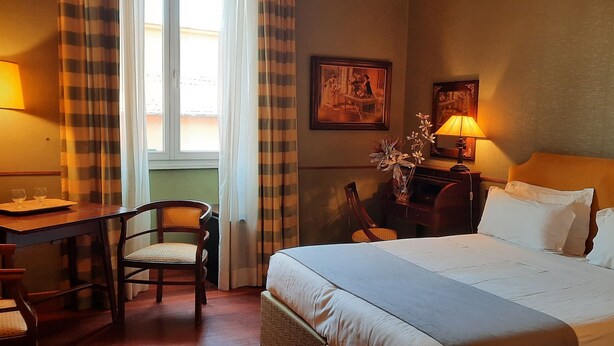 A hotel room with a bed, a desk, a chair, a lamp, and a painting on the wall. - Domus Livia Luxury Suites (Rome)