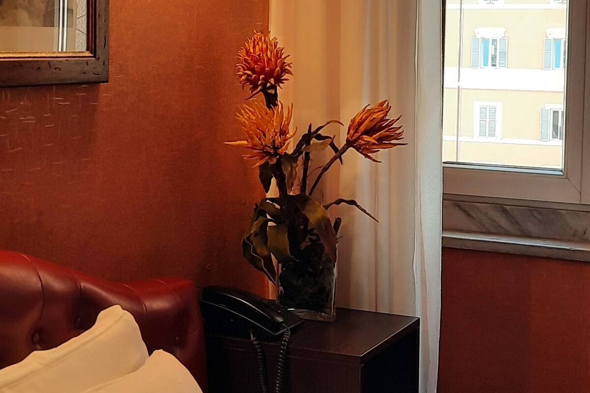 Habitació d'hotel amb un llit, una taula de nit amb un ram de flors, una làmpada penjada a la paret i una finestra amb vista a edificis.