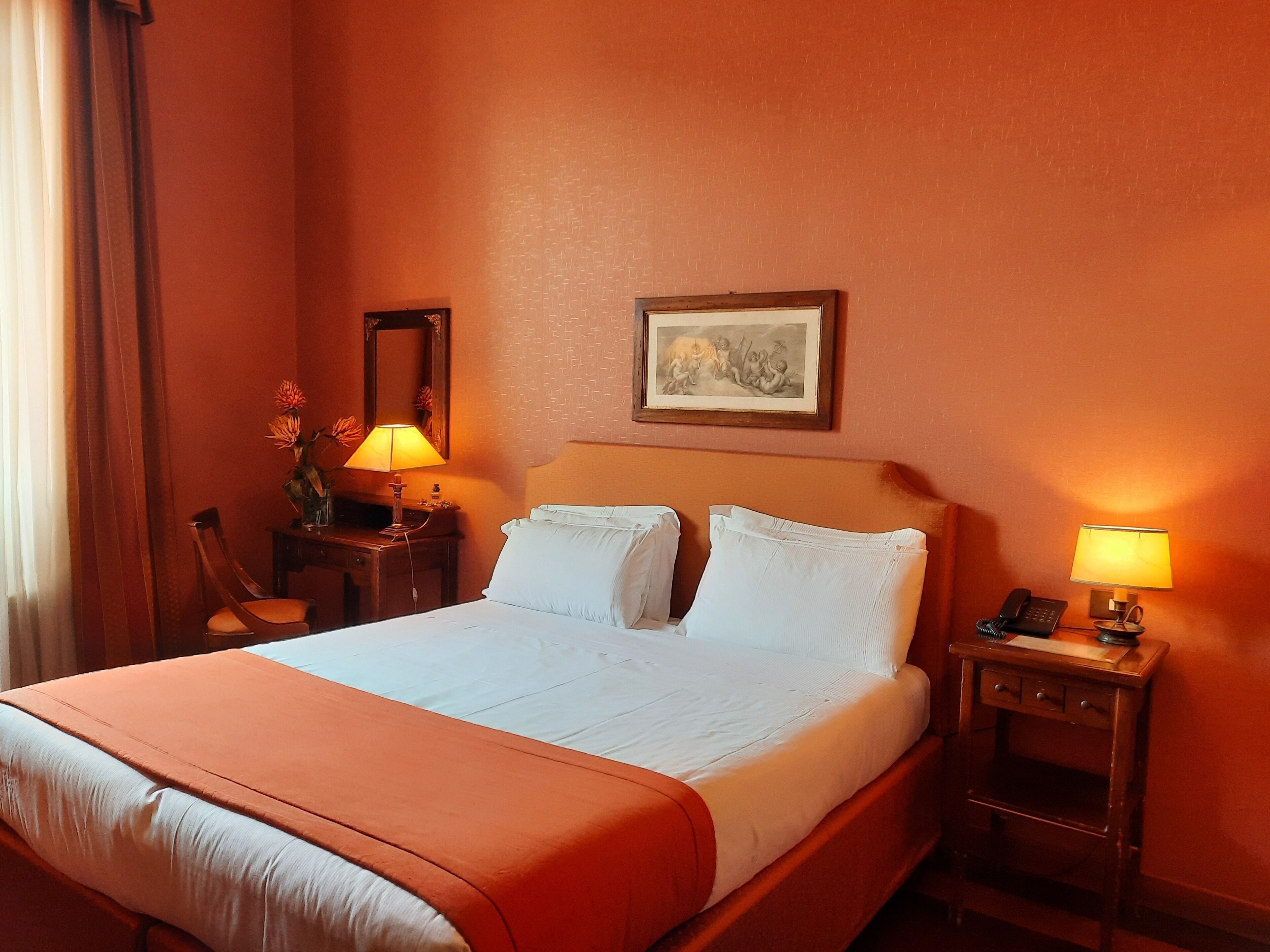 A hotel room with a bed, two bedside tables, a nightstand, a lamp, a mirror, and a painting on the wall.