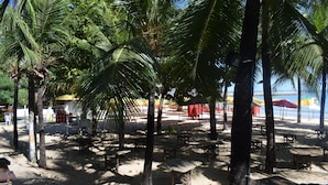 Un café frente a la playa con mesas al aire libre, palmeras y sombrillas.