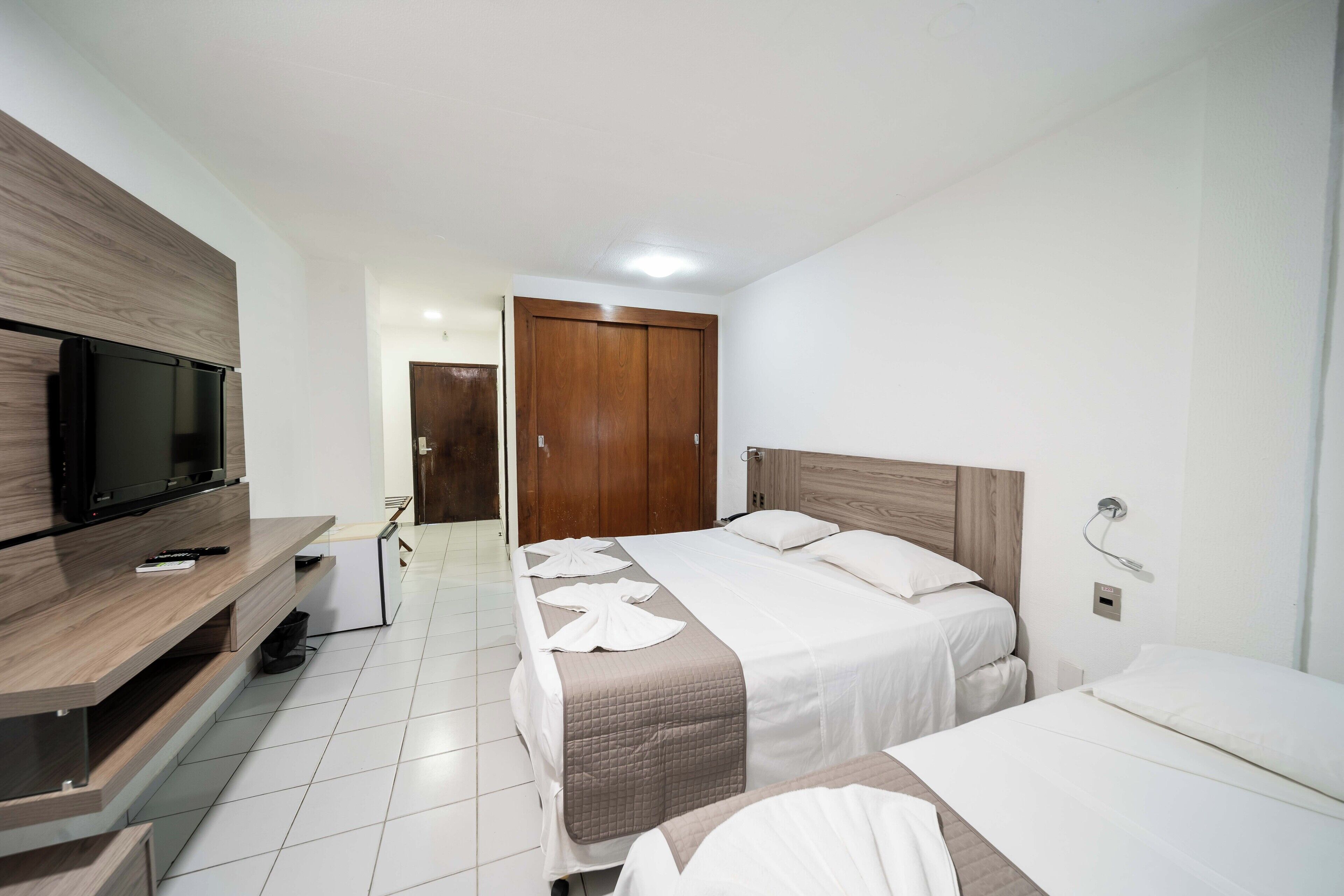 Una habitación de hotel con dos camas, televisión, armario de madera y suelo de baldosas blancas.