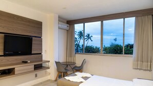 Habitación de hotel con televisión en un estante de madera, una pequeña mesa con dos sillas, una cama con ropa blanca y una ventana que ofrece vistas a palmeras y un edificio.